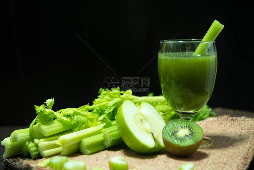 混合汁芹菜和猕猴桃和青苹果在欢迎饮料玻璃和一堆新鲜的芹菜茎放在木桌上图片