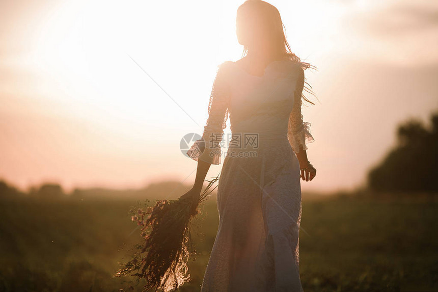 日落时在玉米田上穿着长白色蕾丝裙的年轻女子图片
