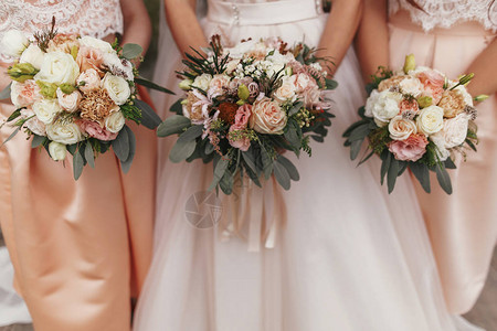 华丽的新娘和美丽的伴娘在现代礼服的背景上拿着时尚的婚礼花束图片