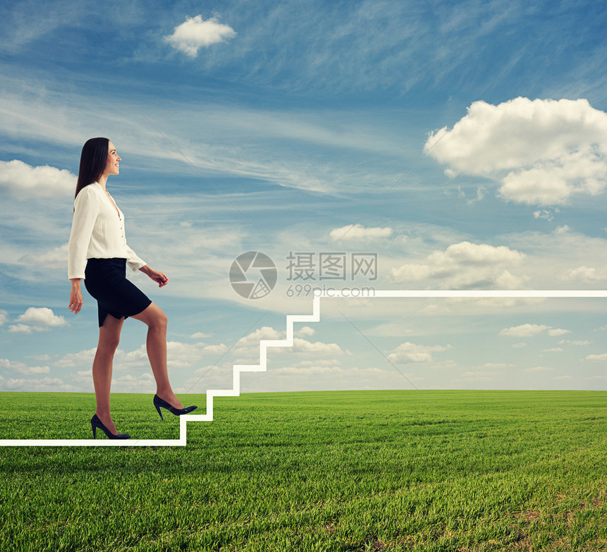 身穿正式服装的微笑女商人在绿色田地和蓝天空上走着白楼梯图片