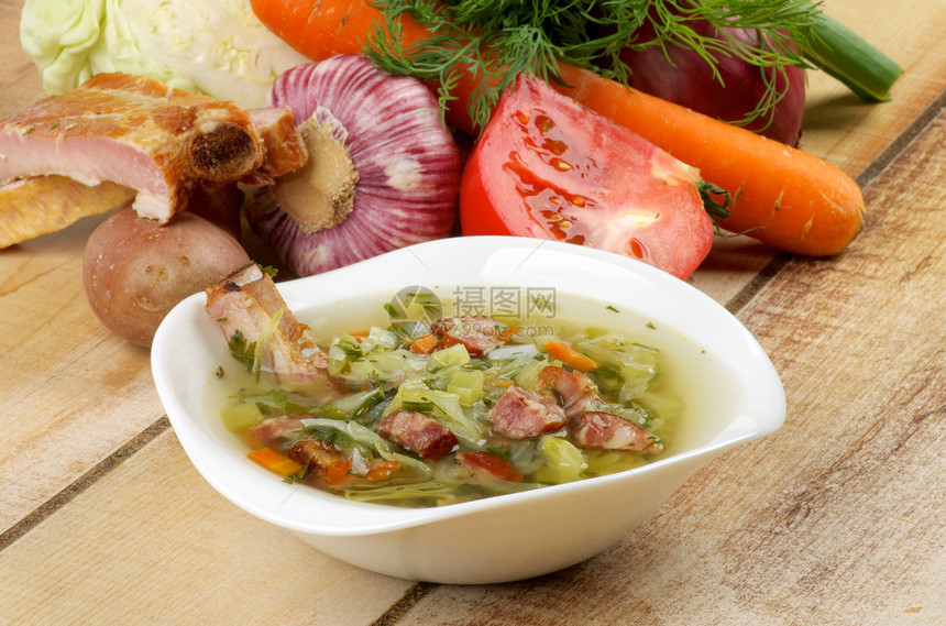 白碗美食蔬菜汤和木制背景上的熏排骨卷心菜西红柿葱韭菜胡萝卜大蒜图片