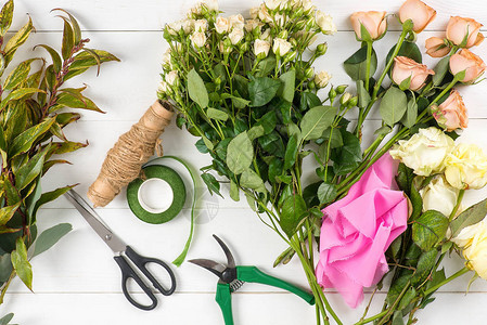 花店从玫瑰和装饰树枝上收集一束花在玫瑰剪刀剪枝机丝带卷和黄麻绳索的白图片