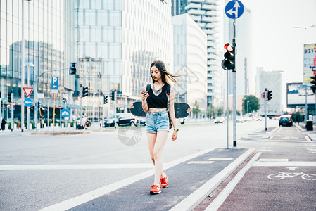年轻美丽的亚洲女子滑冰运动员背着包在城市户外使用智能手机持图片