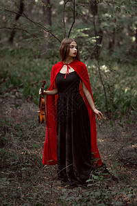 穿着黑裙子和红斗篷的优雅神秘女孩在黑暗的树林背景图片
