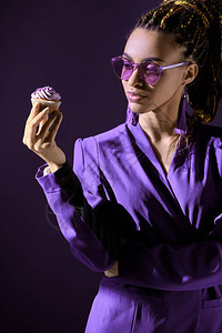 穿着紫外夹克和太阳眼镜的时髦的非裔美国女孩看着纸杯蛋糕孤图片