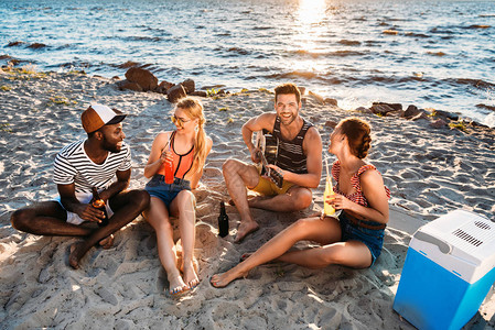 年轻多民族朋友在日落时在沙滩上享受吉他和饮料图片