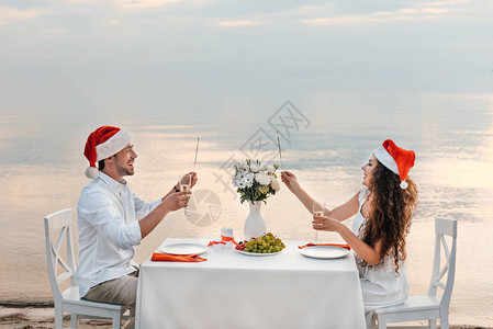 圣诞快乐的一对情侣在圣塔帽子中庆祝圣诞节喝香槟杯子和火图片
