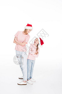 快乐的母亲和女儿戴着圣塔的帽子站着双手交叉看着对方孤立在白边背景图片
