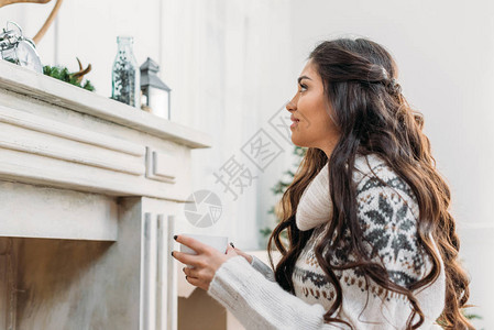 身穿温暖毛衣的美女站在壁炉前喝背景图片
