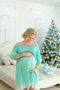 身穿蓝色连衣裙的白种人孕妇抱着肚子站在卧室的圣诞树旁寒假和等图片