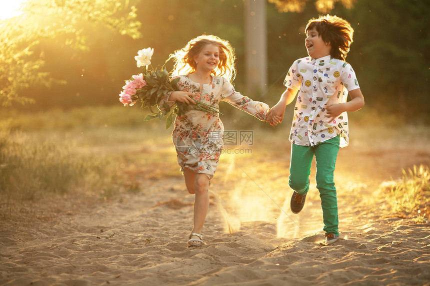 带着花朵的小快乐女孩和男孩在阳光明媚的森林路上跑来图片