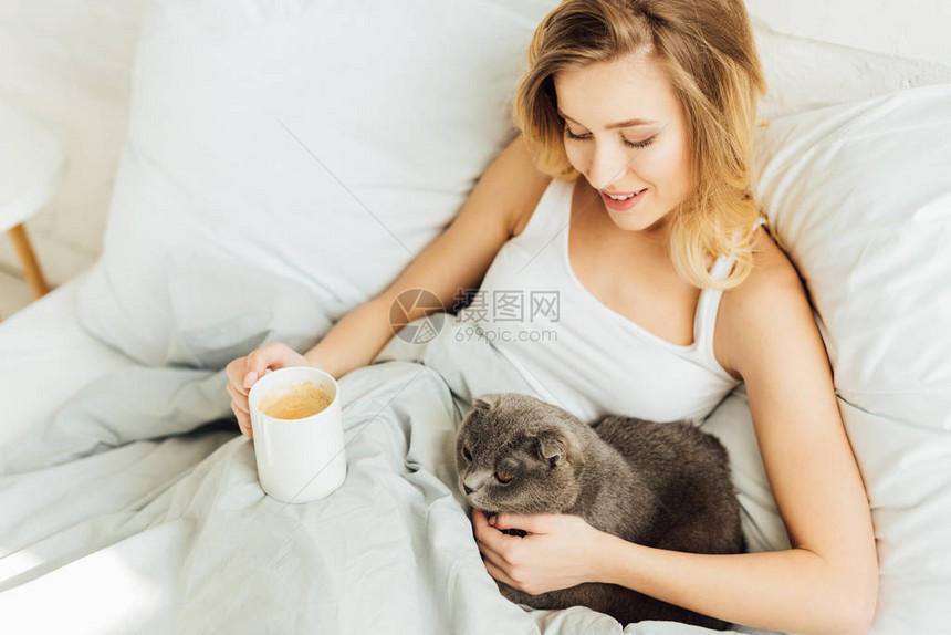 美丽的年轻金发美女躺在床上喝咖啡拥抱苏格图片