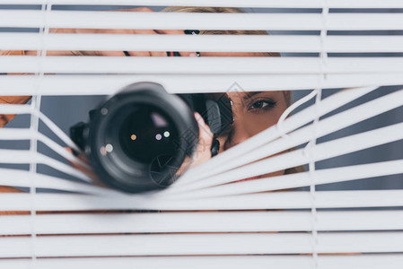 摄影机和年轻妇女拍摄照片并通过百叶窗进行监视的近视图片