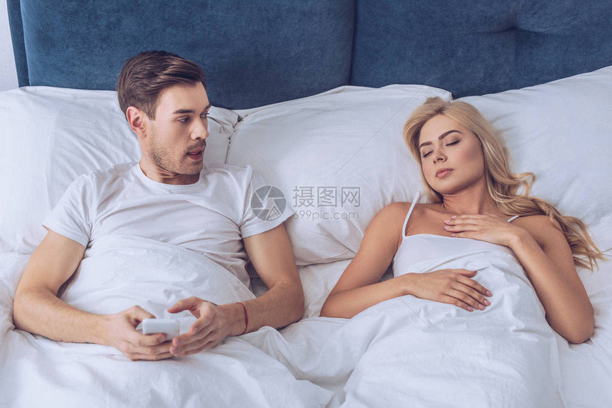 与智能手机一起躺在床上的疑男子和看着睡妻子图片
