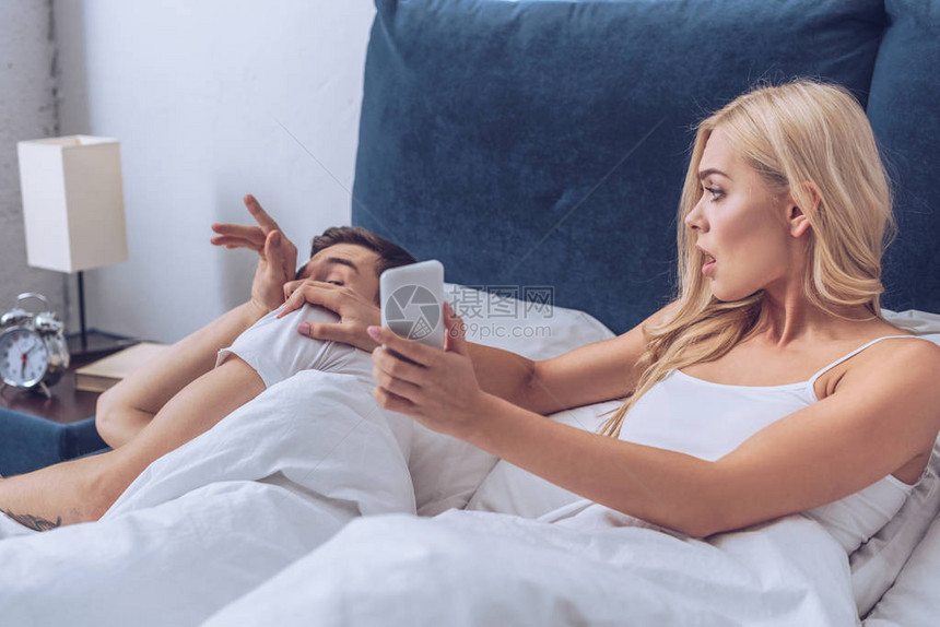 愤怒的老婆用智能手机向躺在床上的困惑丈夫展示了不信任概念图片