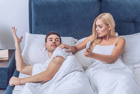 震惊的年轻女子用智能手机向躺在床上的迷乱男友展图片