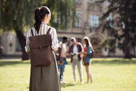 年轻女学生背着包看着同学站在大学附近的草地图片