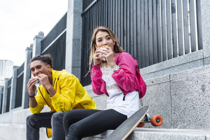 有选择地关注多元文化夫妇在城市街道滑板附近吃汉堡的图片