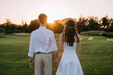 年轻结婚夫妇握着手在日落时一起在公园里站图片