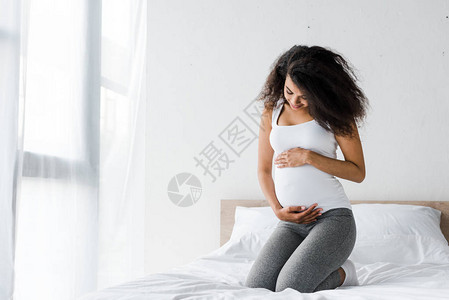 快乐的非洲裔美国孕妇坐在床上时摸着肚子图片