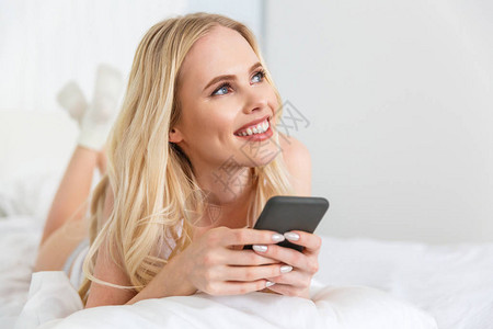 美丽的金发美女使用智能手机躺在床上时图片