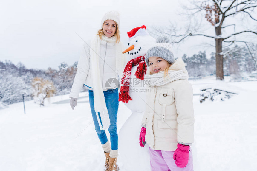 快乐的母亲和女儿在冬季公园与雪人站在一起时图片