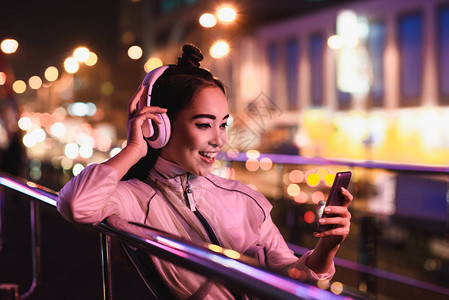 快乐的有吸引力的年轻女孩在街上用智能手机听音乐晚上用夜光收银灯图片