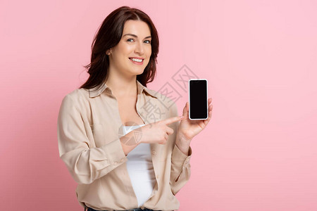 微笑的女人用手指对着智能手机指着粉红背景身体正图片