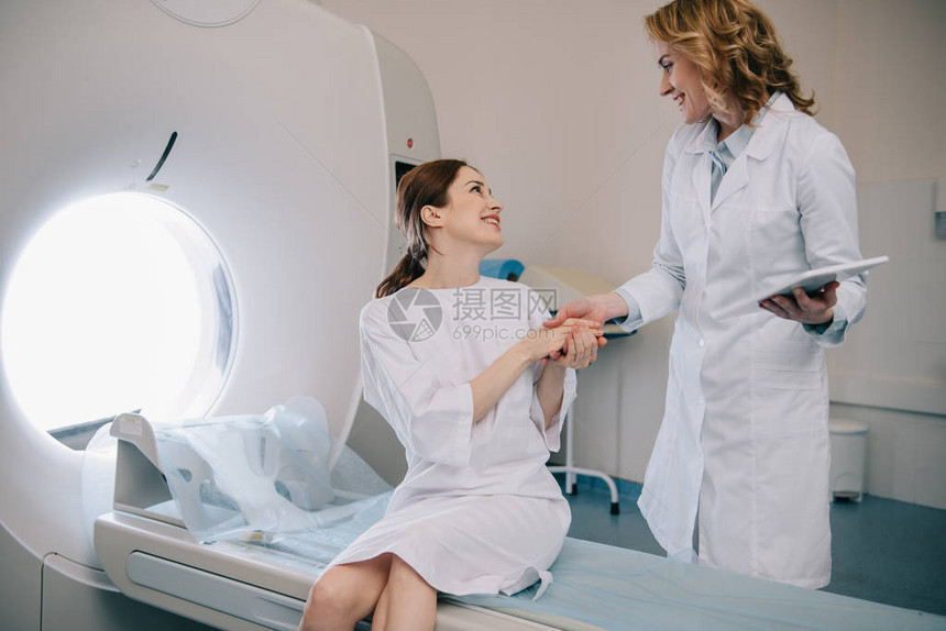 与快乐的女士握手坐在计算成形扫描仪床铺上的放射科医生图片