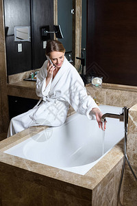 穿着浴袍微笑的年轻女子在酒店坐在浴缸上时用图片