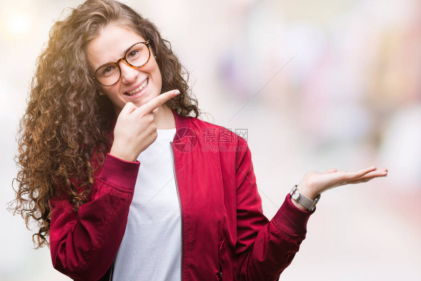 美丽的黑发卷年轻女孩穿着夹克和眼镜在孤立的背景下惊讶并微笑着面对镜头图片