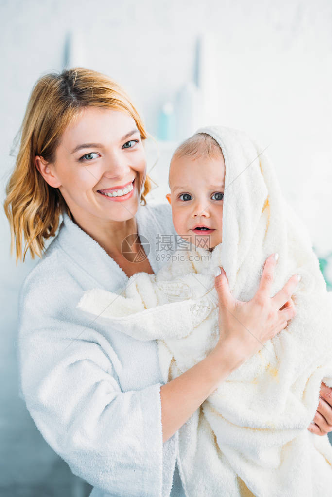 在浴袍中微笑的母亲抱着迷惑的小孩裹着毛巾图片