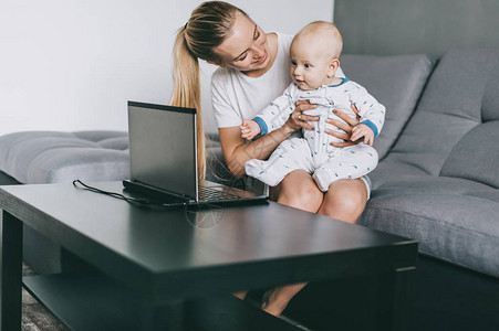 在家使用笔记本电脑时抱着可爱婴儿的年轻母亲图片