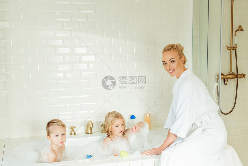 穿着浴袍的美丽的母亲在浴缸中洗孩子时对图片