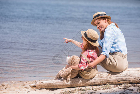 年轻的快乐母亲和女儿与泰迪熊坐在海边时用手指着对图片
