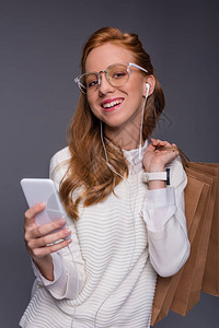 戴着眼镜和白色衣服的微笑时尚红发女孩拿着智能手机和购物袋图片