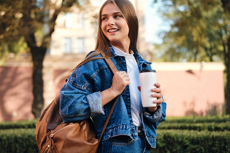 半夏如期而去穿着戴尼姆夹克背包和咖啡的漂亮而愉快的随意学生女孩在大背景