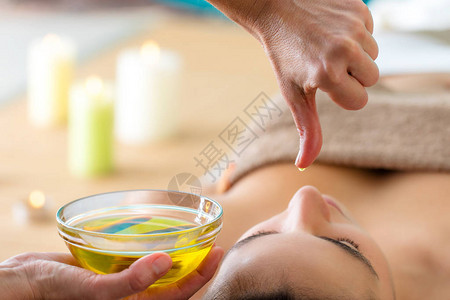 宏观关闭手与油滴在水疗中心的女人头上在阿育吠陀按摩的女人旁边的玻璃碗图片