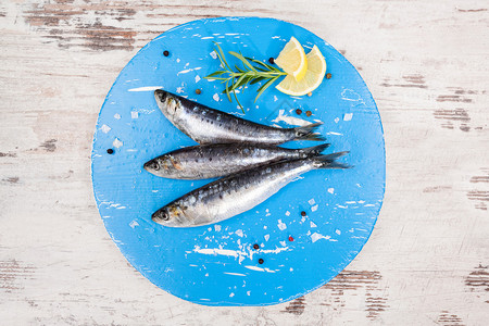 白色木桌上蓝色圆形厨房板上的三条新鲜鳀鱼背景图片