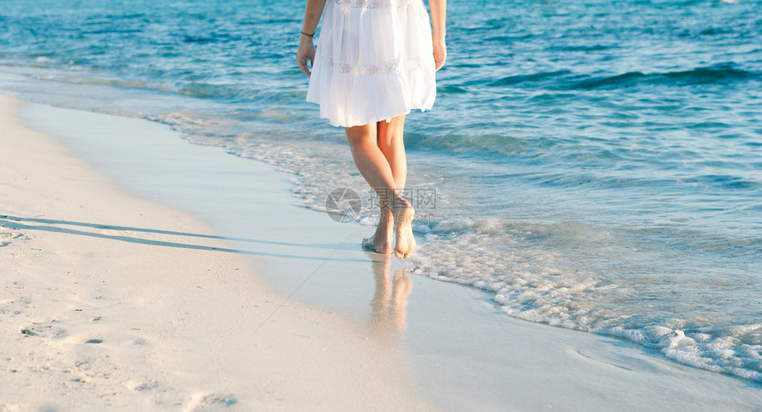 一名年轻女子在日落时沿着白沙滩海岸走下半身图片