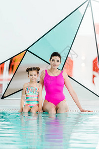 和女儿一起坐在游泳衣上看着泳池附近的照相机她图片