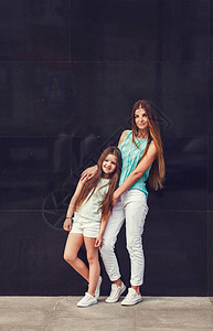 年轻漂亮的妈和她的女儿穿着T恤和牛仔裤靠在水泥墙上摆姿势图片