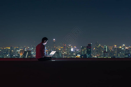 坐在大楼顶部的亚洲商人使用具有城市夜景商业成功和技术理念图片