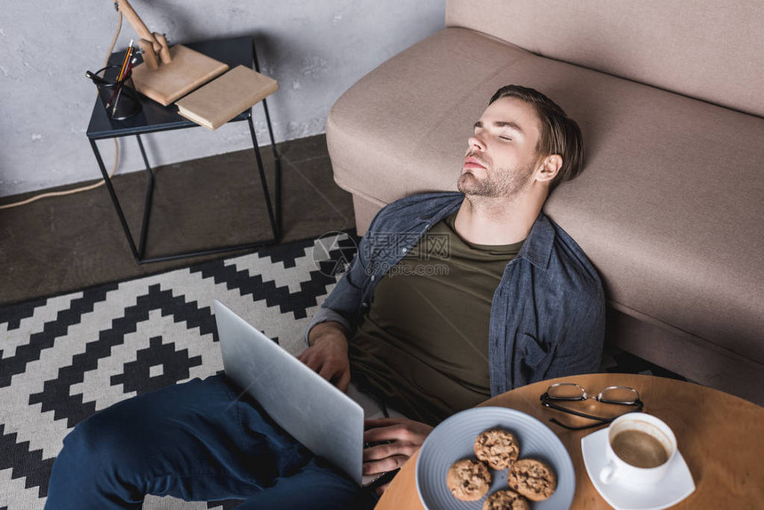 年轻的劳累过度的男人拿着笔记本电脑睡在地板上图片