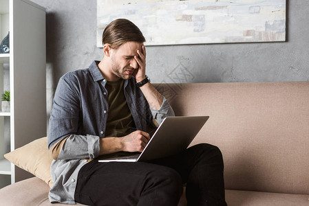 年轻的劳累过度的男人坐在沙发上头疼的笔记本电脑背景图片