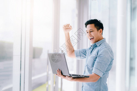快乐兴奋的亚洲男人拿着笔记本电脑举起手臂来庆图片