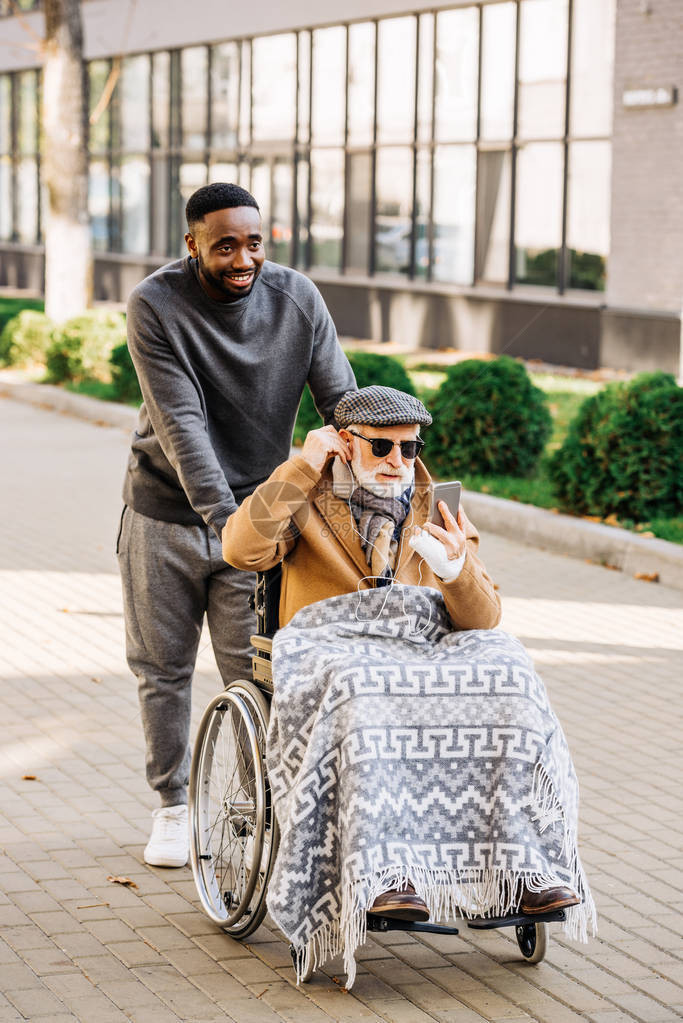 坐在轮椅上的高级残疾人用智能手机和耳机在街上与非洲裔美国骑士图片