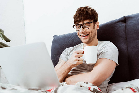 在家用笔记本电脑坐在床上的咖啡杯上笑着喝咖啡的男图片