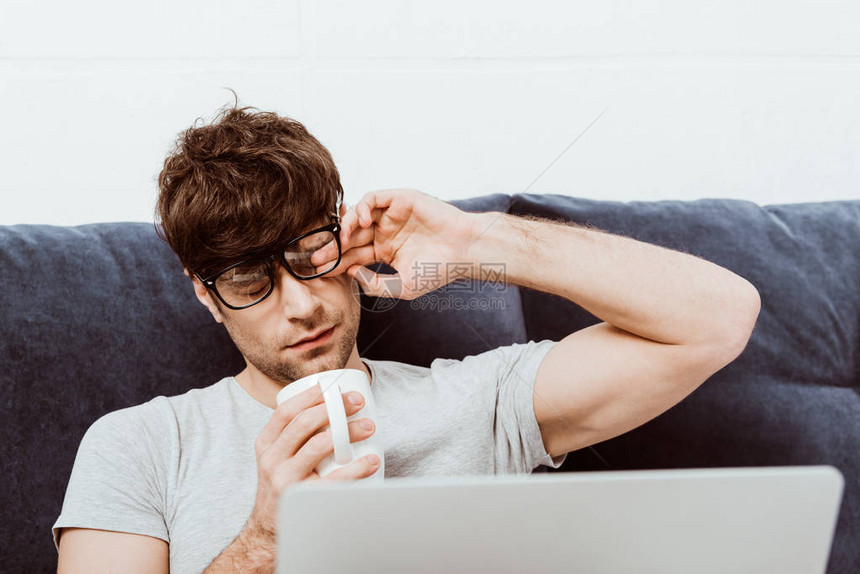 身戴眼镜拿着咖啡杯和坐在家中用笔记本电脑躺在床上的疲累男图片