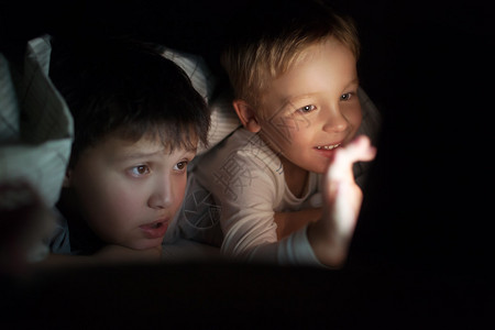 两个男孩晚上躺在床上盖着毯子他们在垫子上看电影或片屏幕在黑暗中图片